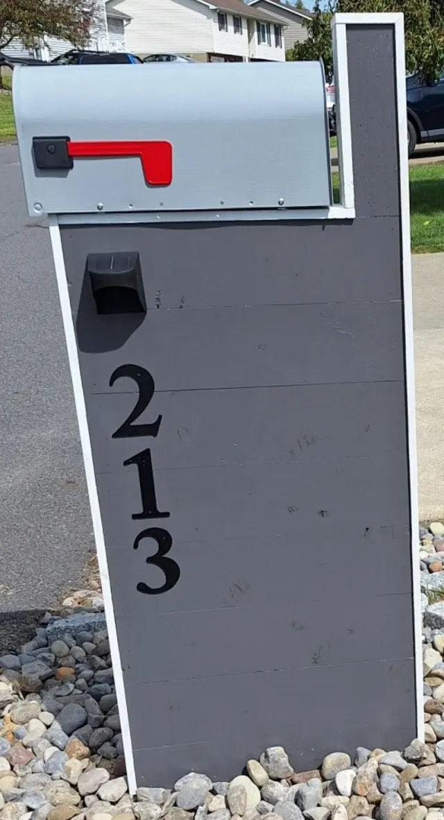 Gray mailbox stand with white trim and black house numbers beside a driveway